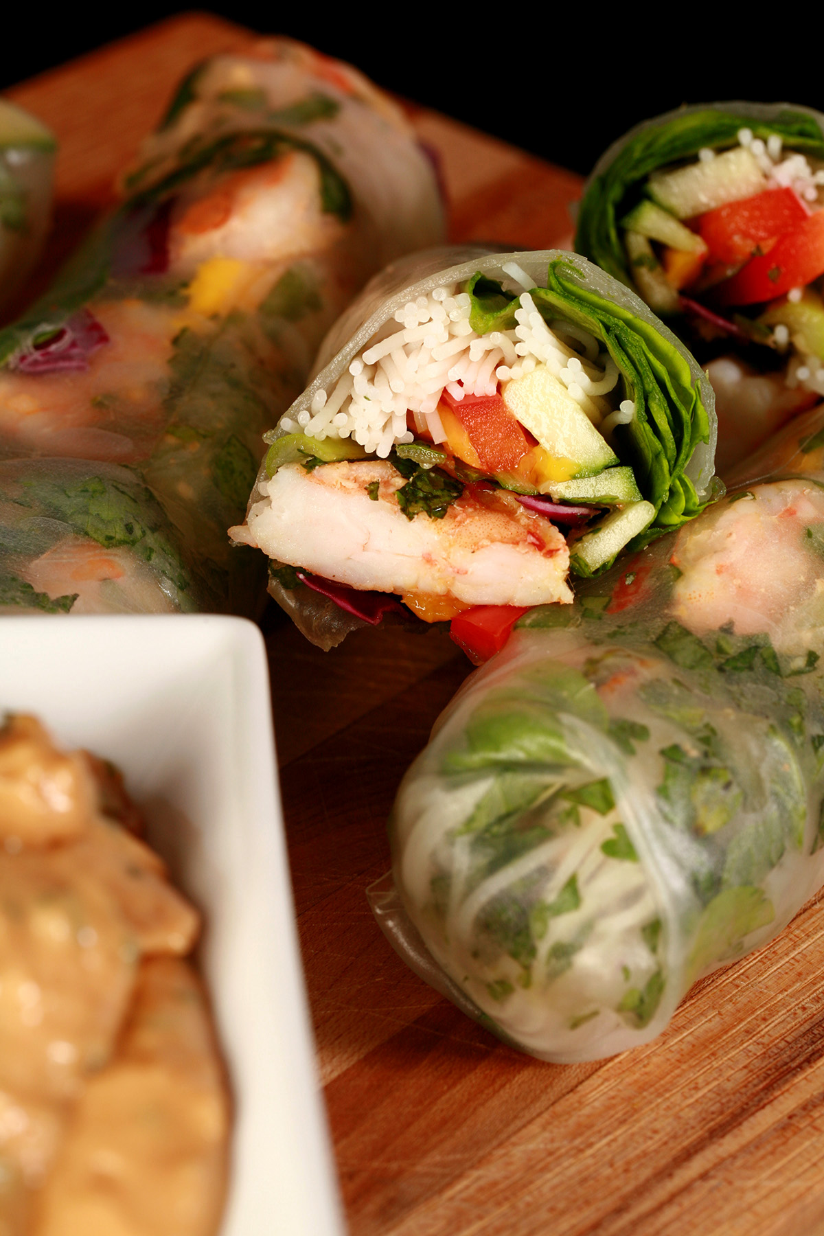 Several mango-shrimp spring rolls on a cutting board, along with a small bowl of peanut sauce.
