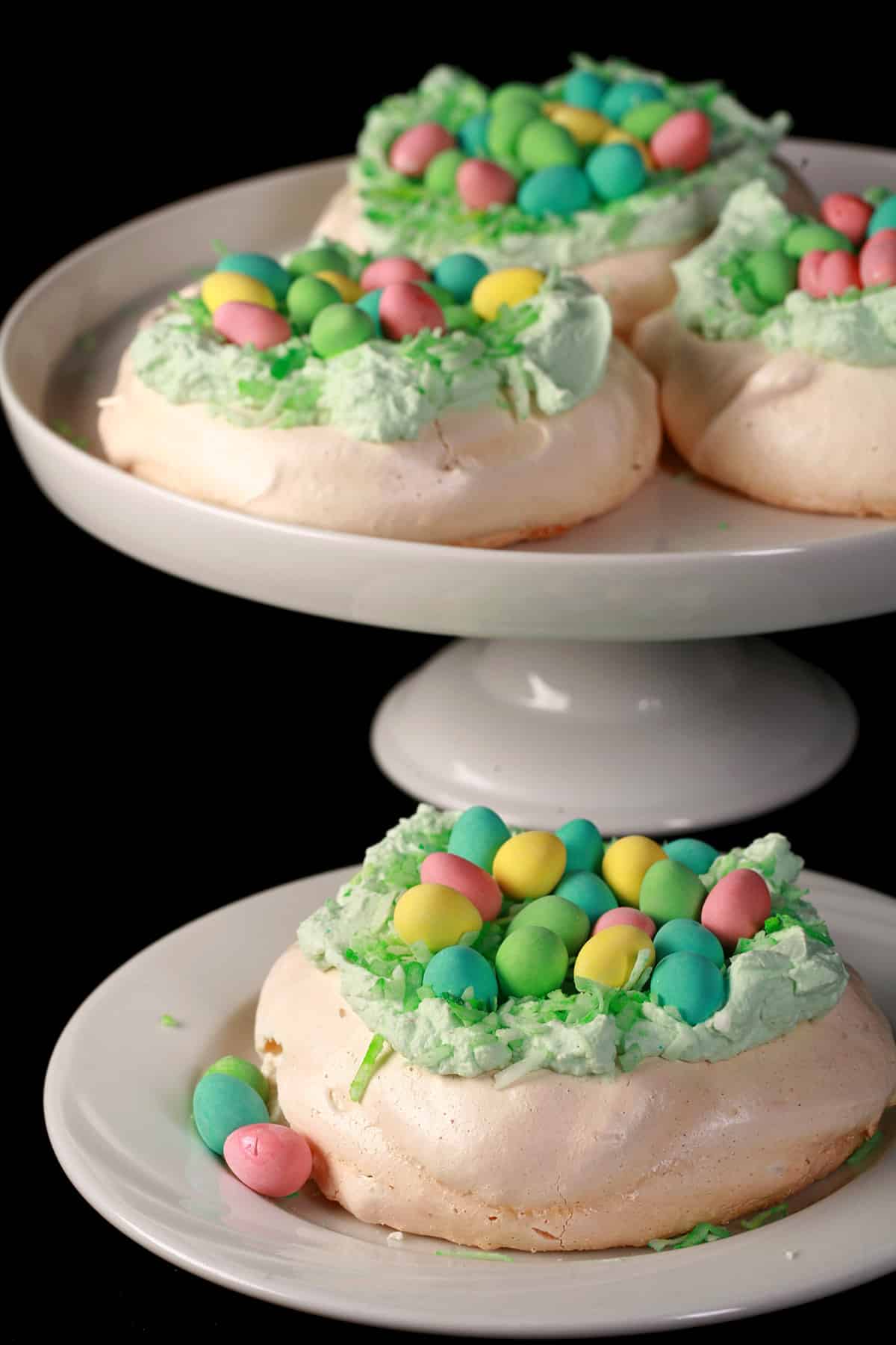 Mini easter pavlovas on a plate and a cake stand. Each have green coconut and cadbury mini eggs on top.