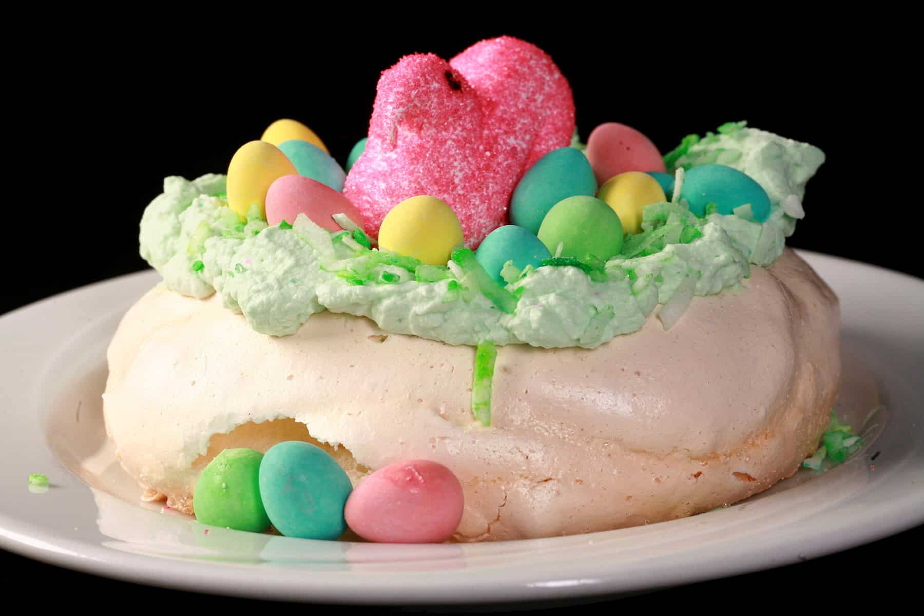 A single mini Easter pavlova, topped with green coconut and cadbury mini eggs and a pink Peeps marshmallow.