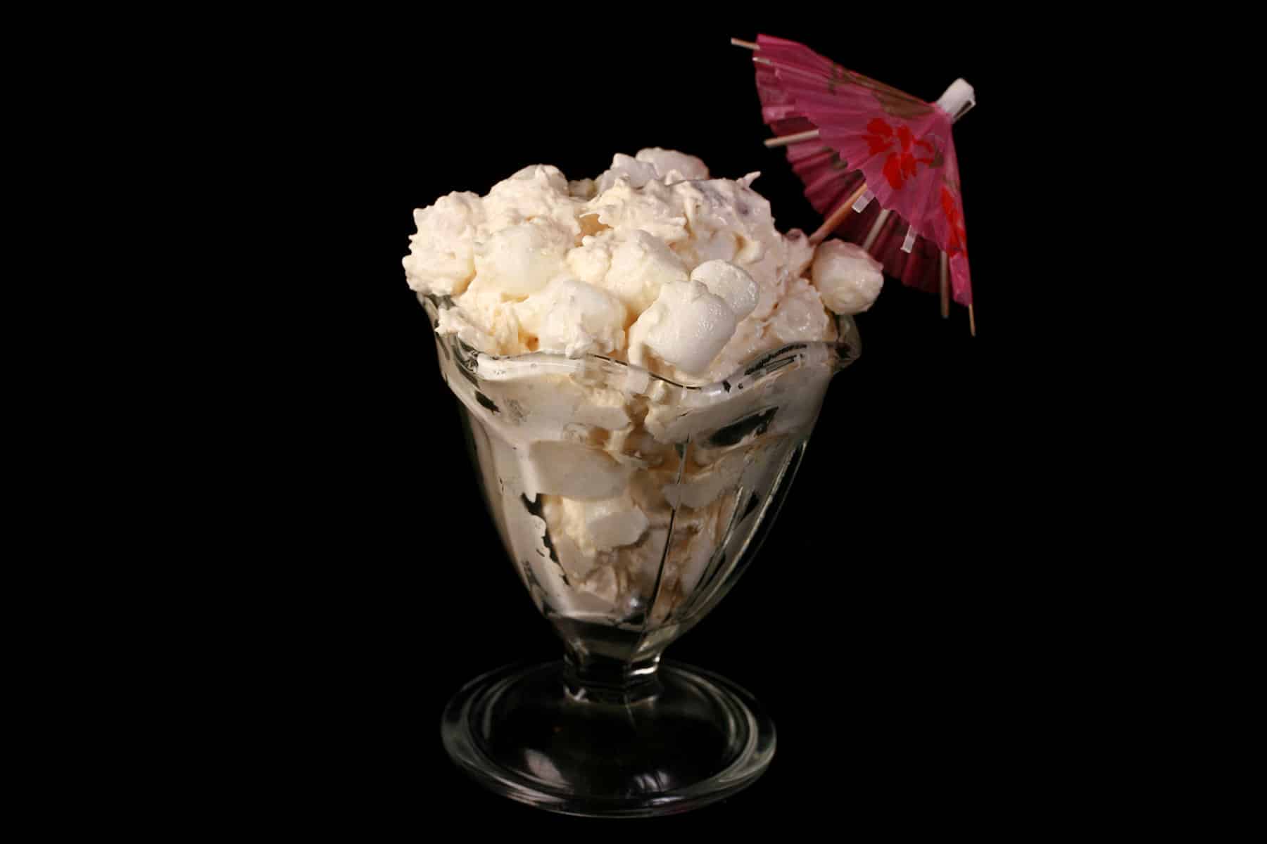 Pina colada marshmallow salad in a tulip glass with a pink cocktail umbrella sticking out.