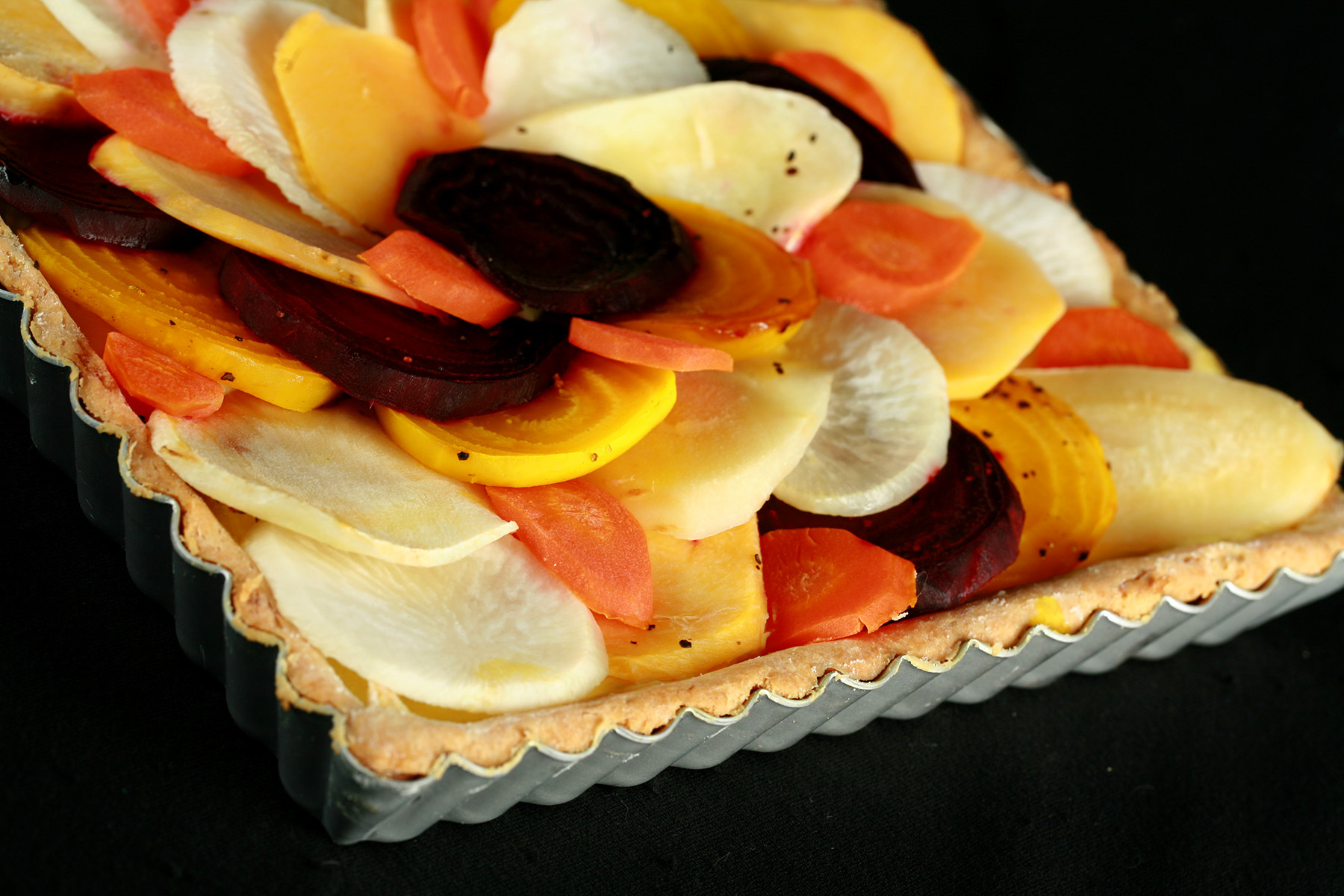 A close up view of a square root tart - a tart cooked in a square pan, with slices of a variety of root veggies arranged on top.