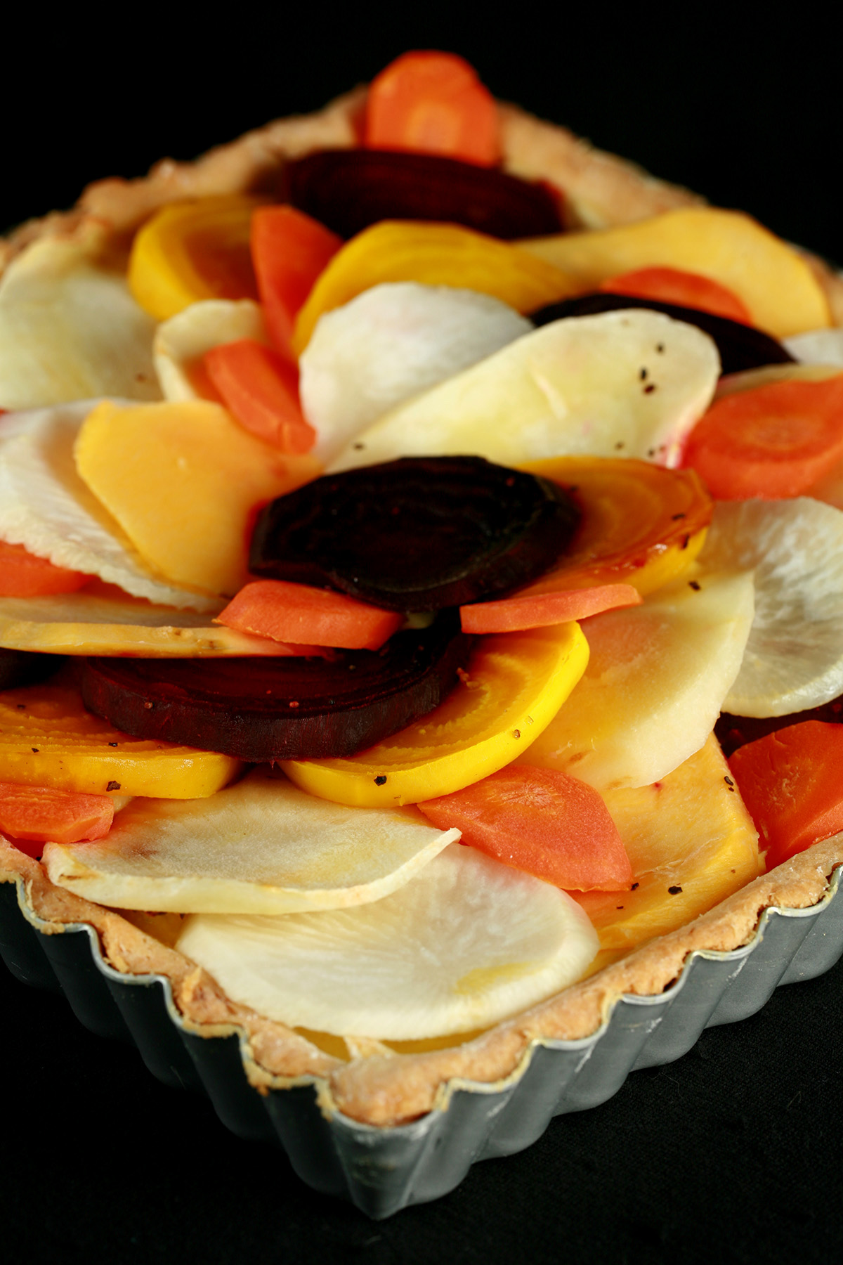 A close up view of a square root tart - a tart cooked in a square pan, with slices of a variety of root veggies arranged on top.